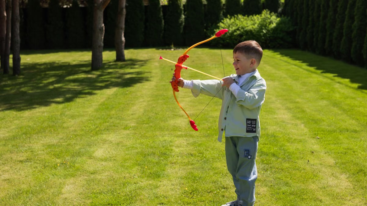 Young boy enjoying a sunny day outside, aiming with a colorful toy bow in a lush garden.
