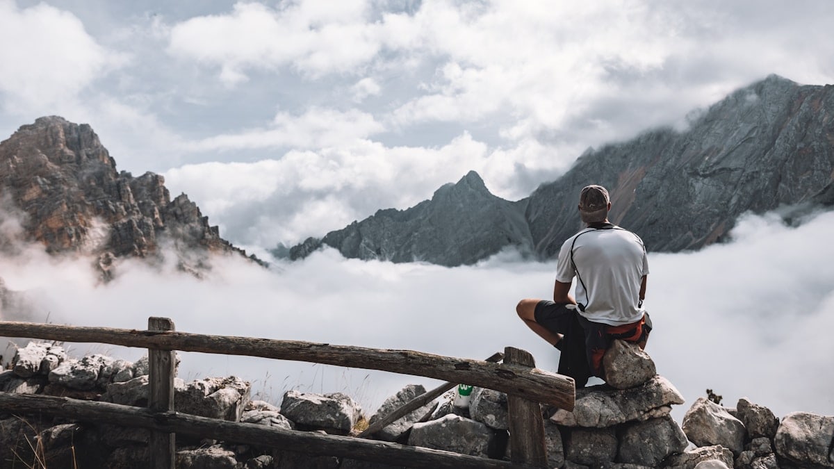 man in white t-shirt sitting on brown wooden fence looking at the mountains during daytime